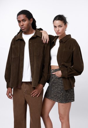 Bruine corduroy jassen met voorkeepockets, gedragen over witte tops. De man combineert het met een paar bruine broeken; de vrouw draagt een spijkerrok met luipaardprint.