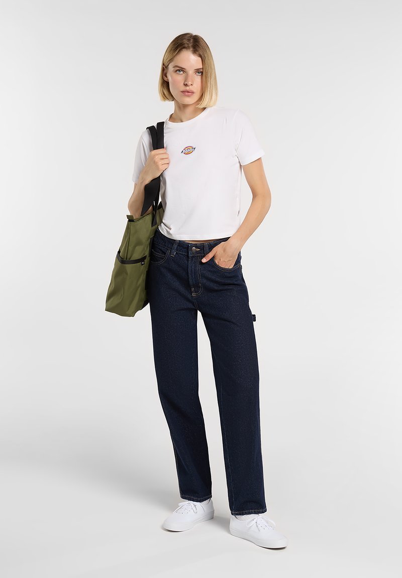 Woman with short blonde hair wearing white logo t-shirt, dark jeans, white sneakers, holding olive green tote bag over shoulder.