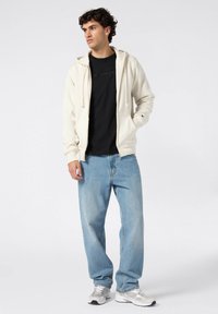 Sweatshirt crème avec des cordons de serrage sur une chemise noire, associé à un jean bleu clair ample et des baskets grises, se tenant sur un fond clair.