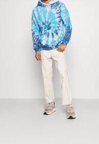 Felpa tie-dye blu e bianca con tasca a marsupio, abbinata a pantaloni color crema e scarpe da ginnastica grigie. Outfit informale con cappuccio regolabile.