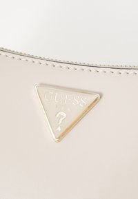 Beige pung i kunstlæder med et trekantet Guess-logo i guldfarve og et præget spørgsmålstegn, fremhævet af en syning langs kanten.