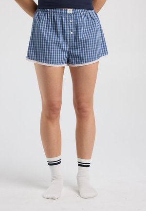 Persoon draagt blauwe geruite boxershorts met knopen en witte sokken met zwarte strepen, staand op een effen witte achtergrond.