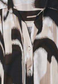 Chemise à col avec motif géométrique abstrait noir, beige et blanc et deux boutons blancs visibles sur la patte de boutonnage avant.