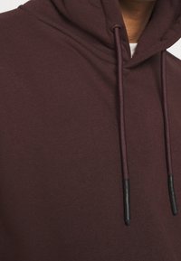 Sudadera burdeos con una textura suave, que cuenta con una capucha ajustable y extremos en negro. Vista en primer plano que enfatiza los detalles de la tela y el diseño.