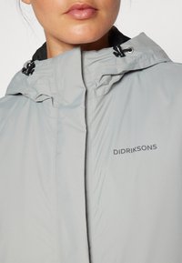 Veste imperméable gris clair avec un col montant, une capuche à cordon réglable et un logo "DIDRIKSONS" sur la poitrine. Texture lisse.