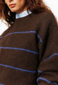 Pull marron en tissu doux et texturé, avec des rayures horizontales bleues. Encolure ronde et coupe ample, mettant en valeur un design décontracté.