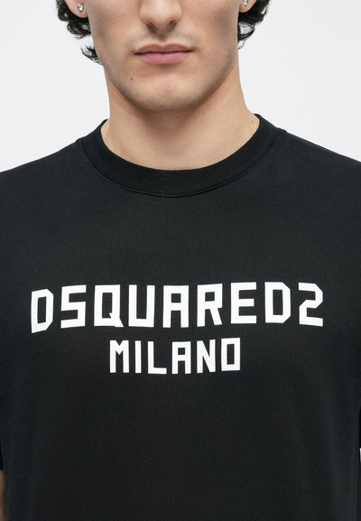 Tuvplāns ar personu melnā T-kreklā ar baltu uzrakstu "DSQUARED2 MILANO" krūšu rajonā, redzama daļa no sejas un kakla.