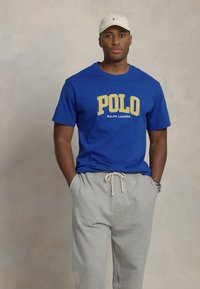 Blue cotton t-shirt with yellow "POLO RALPH LAUREN" text, paired with light gray sweatpants. Textured fabric, relaxed fit.