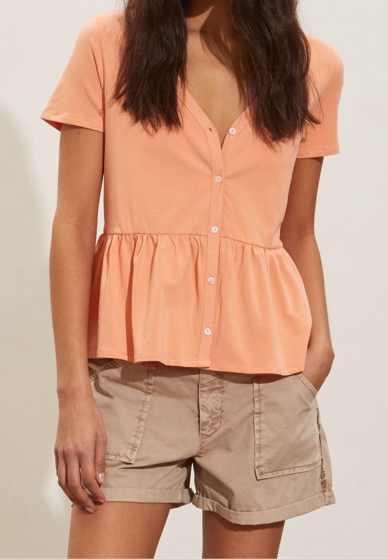Top pesco a maniche corte con chiusura a bottoni frontale, scollatura a V e orlo arricciato. Abbinato a pantaloni corti beige con tasche laterali.