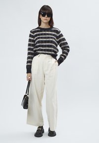 Schwarz-weißer gestreifter Strickpullover, cremefarbene weit geschnittene Hose, schwarze klobige Schuhe mit goldenen Akzenten und eine kleine schwarze Handtasche.