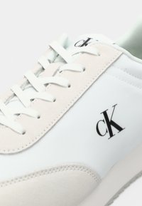 Close-up van een witte sneaker met beige suede accenten, witte veters en een zwart "cK" logo aan de zijkant en op de tong.