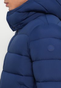 Marineblå jakke med puff gør med quiltede sømme, høj krave og et logo-patch på den øverste arm. Materialet ser glat og isoleret ud.