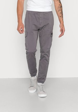 Mann trägt graue Cargo-Hose mit Seitentaschen, weiße High-Top-Sneaker und ein weißes T-Shirt, steht vor einem einfarbigen Hintergrund.