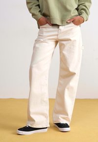Light beige wide-leg pants made of cotton, featuring front pockets, a relaxed fit, and stitched detailing, paired with black sneakers.