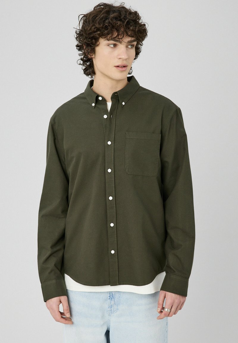Chemise boutonnée à manches longues de couleur vert foncé avec un col, une poche avant, des boutons blancs et un ourlet arrondi, portée par-dessus un T-shirt blanc.