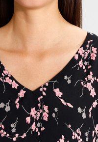 Vêtement noir avec un col en V, présentant des motifs floraux roses et gris. Texture lisse et tissu léger. Vue en gros plan du col.