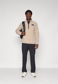 Pullover in fleece beige con zip a un quarto, dettagli neri e pantaloni blu navy. La persona indossa uno zaino nero e sneakers bianche.