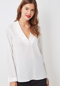 Blusa blanca de manga larga con escote en V y ribete de satén. Tejido suave y ligero con un ajuste relajado, combinada con una falda negra texturizada.