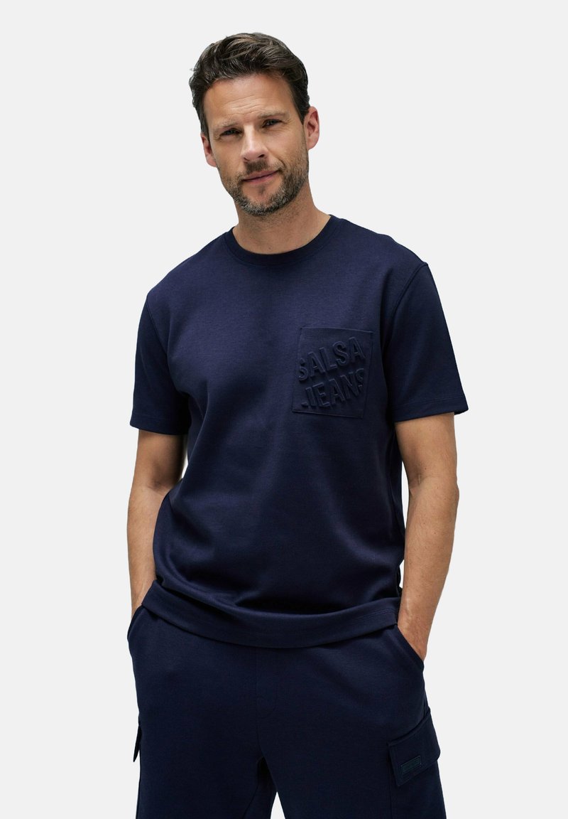 Camisa de algodón azul marino con cuello redondo, mangas cortas y un bolsillo frontal con el logotipo "SALSA JEANS" en relieve. Textura suave.