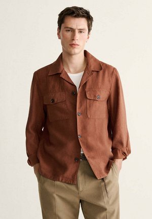 Junger Mann mit Händen in den Taschen, der ein rostbraunes Hemd mit Knöpfen über einem weißen T-Shirt und beigen Hosen vor einem schlichten Hintergrund trägt.