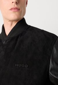 Black bomber jacket with a suede body, leather sleeves, ribbed collar, and debossed "HUGO" logo on the front.