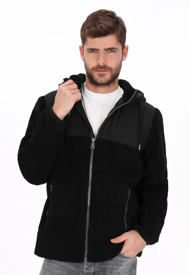 Chaqueta de forro polar negra con un panel superior mate en negro contrastante, cierre de cremallera, capucha ajustada y bolsillos laterales. Tejido suave y texturizado.