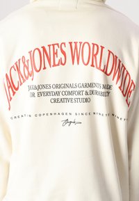 Hoodie couleur crème avec un texte rouge et noir au dos, incluant "JACK&JONES WORLDWIDE" et d'autres détails de marque. Matière douce.