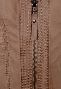 Detail béžového koženého kabátu se zipem, vybavený kovovým zipem, dvojitým prošitím a texturovaným povrchem podél okrajů a švů.