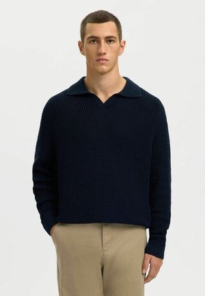 Mornarsko plavi pleteni pulover s V-izrezom i širokim ovratnikom, s rebrastim manšetama i rubom, nošen preko bež hlača.