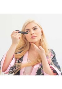 Czarny masażer do twarzy trzymany w dłoni, używany na policzku. Modelka ma na sobie kwiecistą szlafrok z różowymi akcentami, prezentując długie blond włosy i neutralny makijaż.