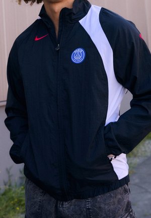 Personne portant une veste noire et blanche du Paris Saint-Germain avec un logo Nike rouge, les mains dans les poches, debout en plein air.