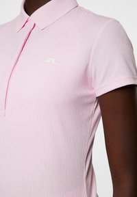 Jasnoróżowa koszulka polo z fakturowanej tkaniny, z klasycznym kołnierzykiem i białym logo na piersi. Krótkie rękawy i zapięcie na guziki.