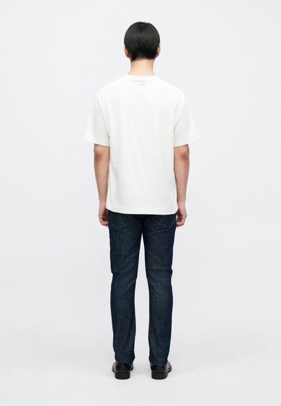 Λευκό βαμβακερό t-shirt με στρογγυλή λαιμόκοψη, κοντά μανίκια και ίσια ραφή στο κάτω μέρος, συνδυασμένο με σκούρο μπλε τζιν και μαύρα παπούτσια.