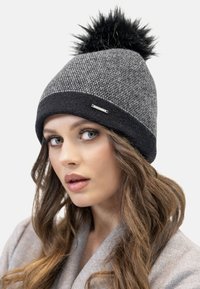 Gorro de punto gris y negro con un patrón texturizado, rematado con un pompón de piel sintética negra; etiqueta de acento plateado en la banda.