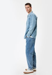 Giacca in denim azzurro chiaro con vestibilità rilassata e maniche lunghe, abbinata a jeans a gamba larga dello stesso colore e sneakers bianche con dettagli marroni.
