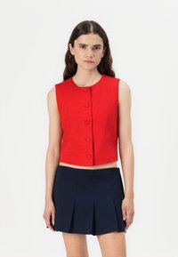 Top corto sin mangas de color rojo con escote redondo, que cuenta con cinco botones visibles en la parte delantera y una silueta ajustada. Combinado con una falda plisada de color azul marino.
