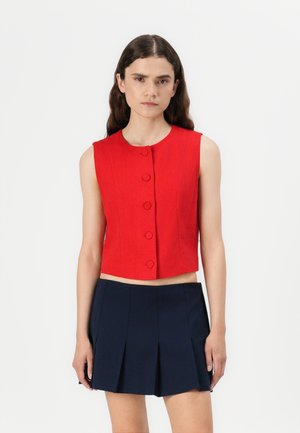 Rød, ermeløs cropped topp med rund hals, med fem synlige knapper foran og en tettsittende silhuett. Bært med et marineblått plisserte skjørt.