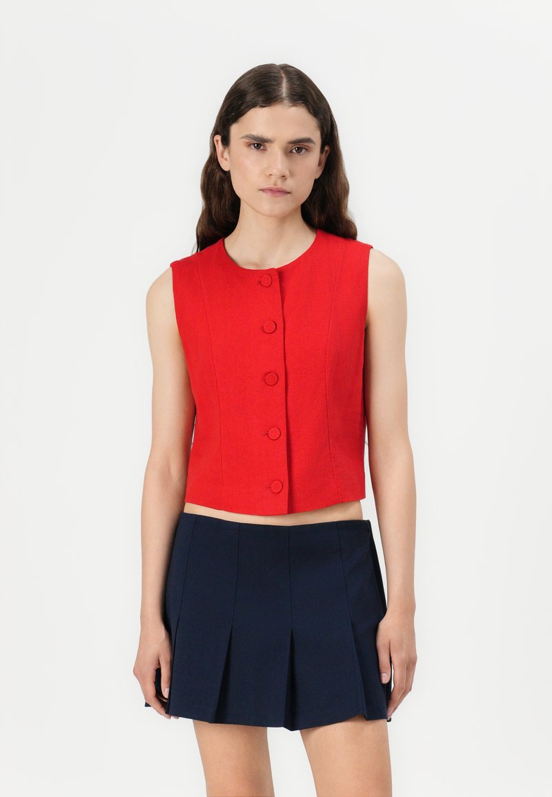 Top corto sin mangas de color rojo con escote redondo, que cuenta con cinco botones visibles en la parte delantera y una silueta ajustada. Combinado con una falda plisada de color azul marino.