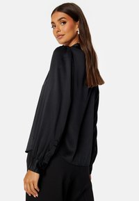 Blusa in satin nera con maniche lunghe a sbuffo, colletto alto e orlo leggermente curvato. Texture liscia con una leggera lucentezza.