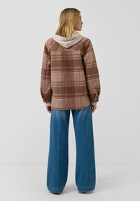 Brązowa kraciasta koszula z beżowymi akcentami, z kapturem, długimi rękawami i luźnym krojem. W zestawie z niebieskimi dżinsami o szerokich nogawkach.