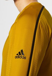 Maglietta sportiva gialla a maniche lunghe con dettagli testurizzati, caratterizzata da logo adidas nero e strisce laterali nere. Design casual, silhouette aderente.
