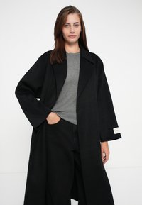 Black oversized coat with wide sleeves, made of textured fabric. Worn over a grey shirt, features a label on the sleeve and deep pockets.