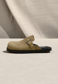 Sabot marron clair à enfiler avec bout arrondi, bride ajustable à boucle et semelle noire coussinée sur une surface texturée claire.