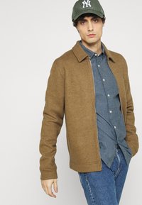Bruine sweaterjas met een kraag, gedragen over een blauwe blouse. Het model draagt ook een groene pet, met een neutrale achtergrond.