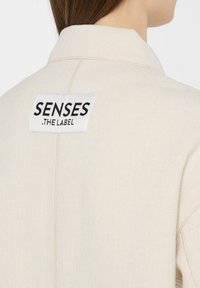 Beige Jacke aus strukturiertem Stoff mit einem schwarz-weißen Etikett auf der Rückseite, in der Nähe des Kragens, das "SENSES .THE LABEL" liest.