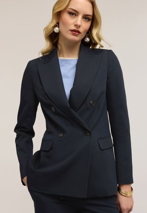 Blonde Frau trägt marineblauen, doppelreihigen Blazer, hellblaues Oberteil, Perlenohrringe und goldene Armreifen, steht vor neutralem Hintergrund.