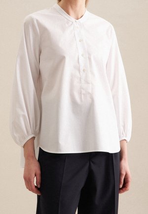 Osoba ubrana w luźną białą bluzkę z długim rękawem, z kołnierzykiem typu stójka i czterema guzikami, w połączeniu z czarnymi, dopasowanymi spodniami.