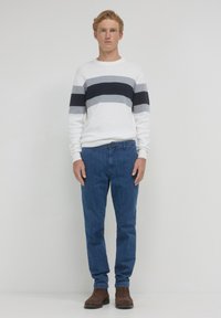 Giovane uomo dai capelli rossi che indossa un maglione bianco con strisce grigie e blu scuro, jeans blu e stivali marroni, in piedi su uno sfondo bianco semplice.