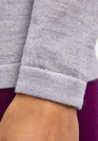 Polsino di maglione lavorato a maglia grigio chiaro con una finitura texturizzata. Il polsino è arrotolato, rivelando un bordo liscio. La mano poggia su una superficie di tessuto viola.