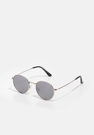 Ochelari de soare rotunzi cu lentile gri închis, ramă subțire din metal argintiu și silenții din cauciuc negru. Design simplu, fără modele sau accente.
